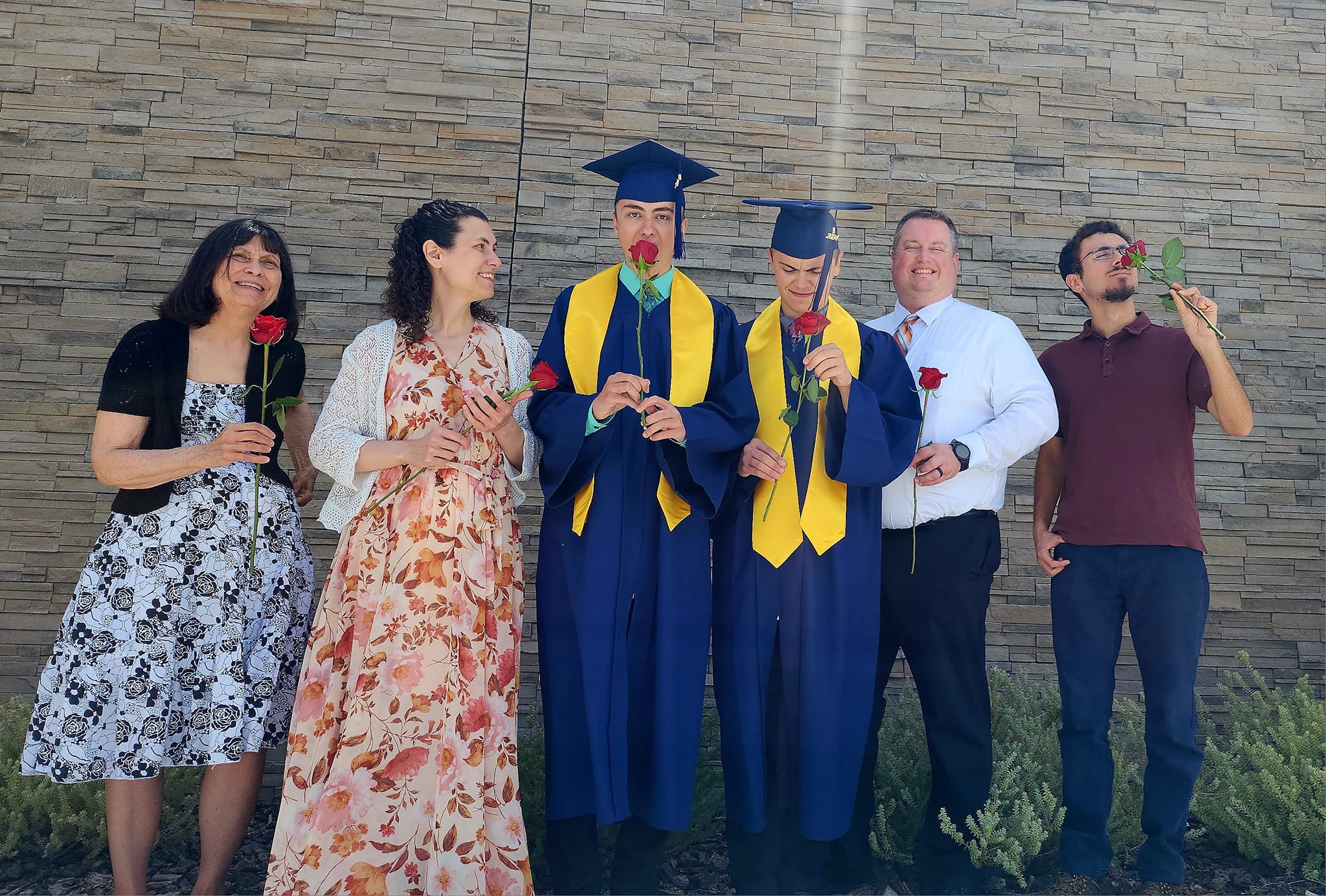 Graduates in caps and gowns hold roses, surrounded by family in a celebratory pose against a stone wall, marking a graduation milestone.