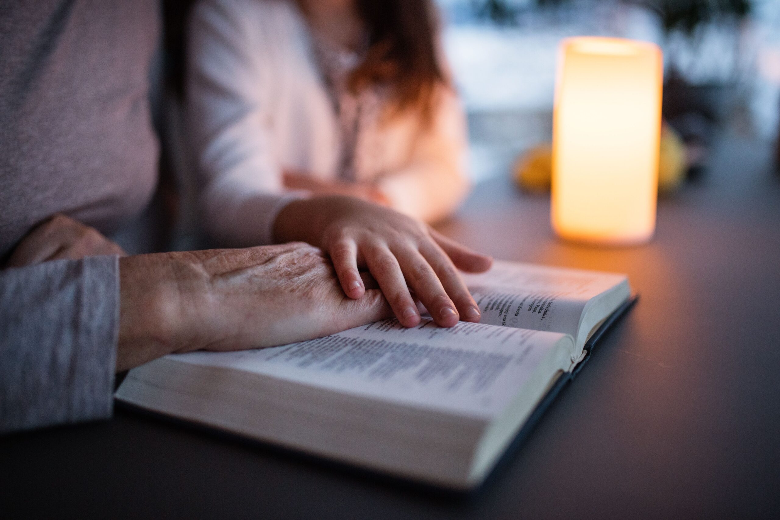 An adult's hand gently rests on an open book, while a child's hand is placed on top. A warm candlelight casts a soft glow, creating a cozy reading moment.